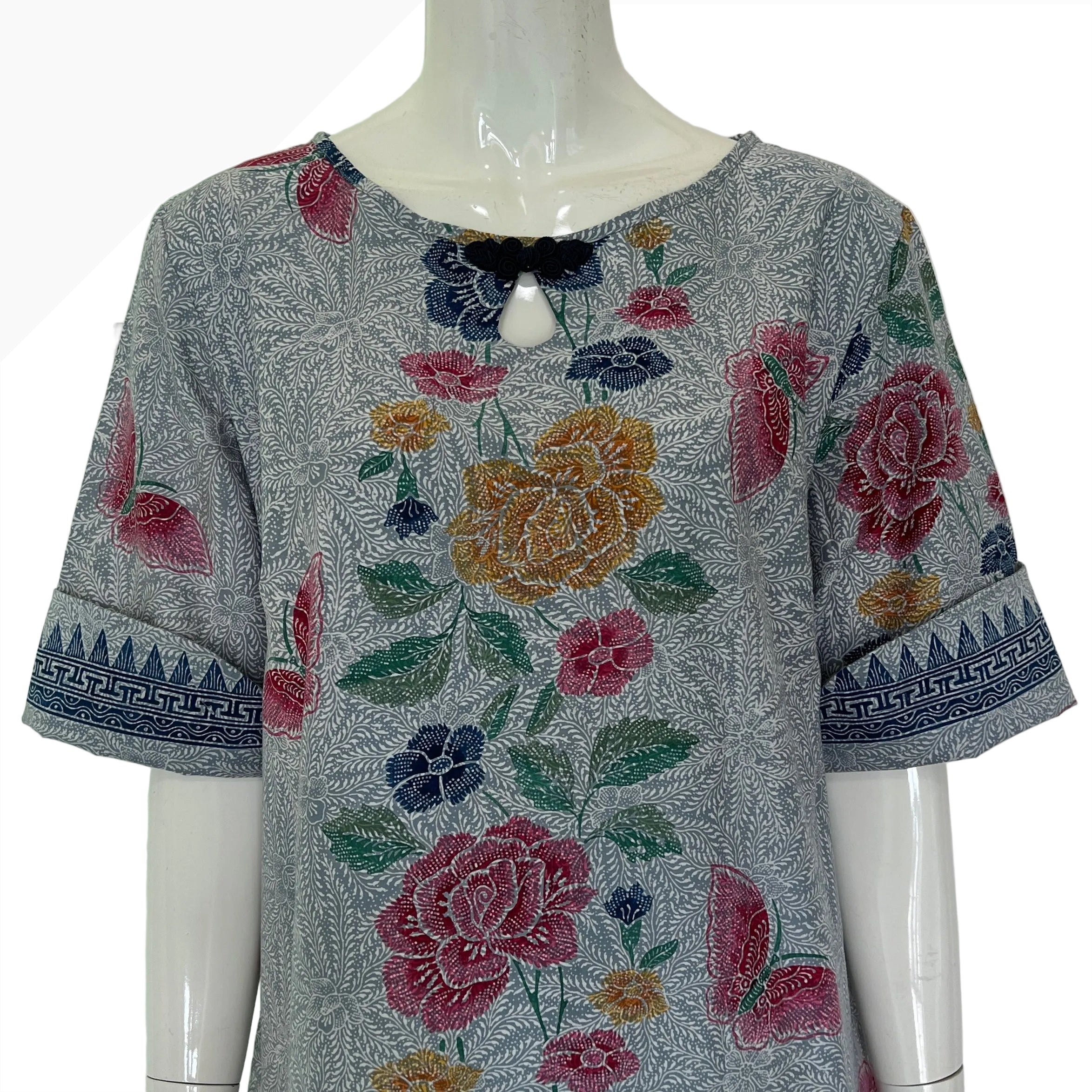 A short-sleeve gray patterned blouse displayed on a mannequin, featuring colorful floral embroidery or printed motifs in pink, yellow, green, and blue, with a geometric blue border on the sleeve cuffs.