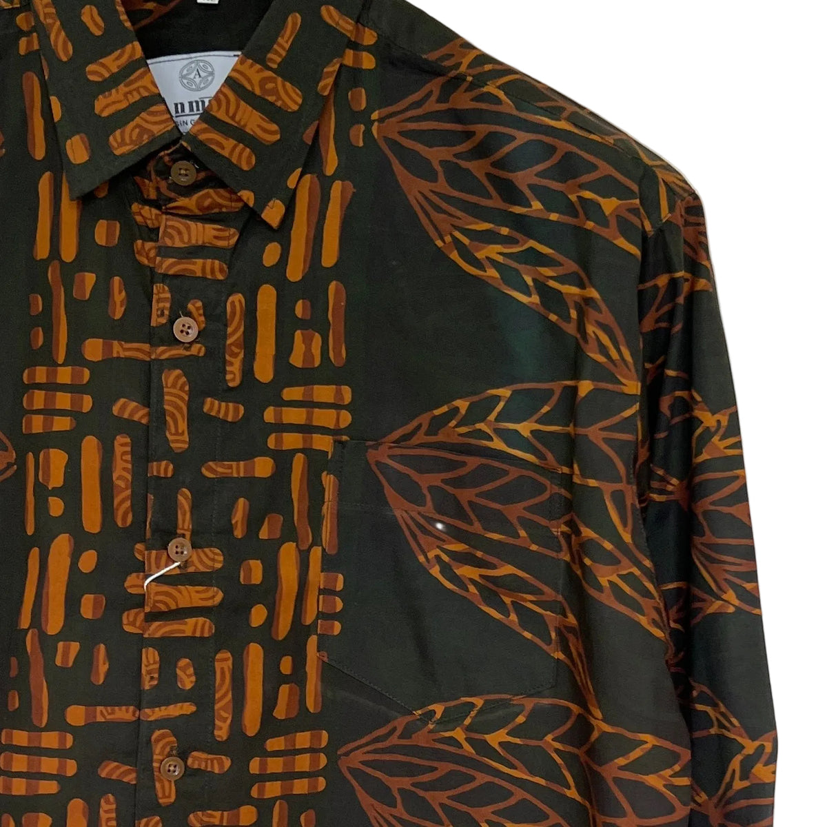 A close-up of a long-sleeve button-up shirt with a dark charcoal base featuring a split design: one side has a pattern of orange-brown vertical bars, and the other features large orange-brown leaf motifs. The shirt has brown buttons and a visible inner collar label with the brand name 'BLOOM'.